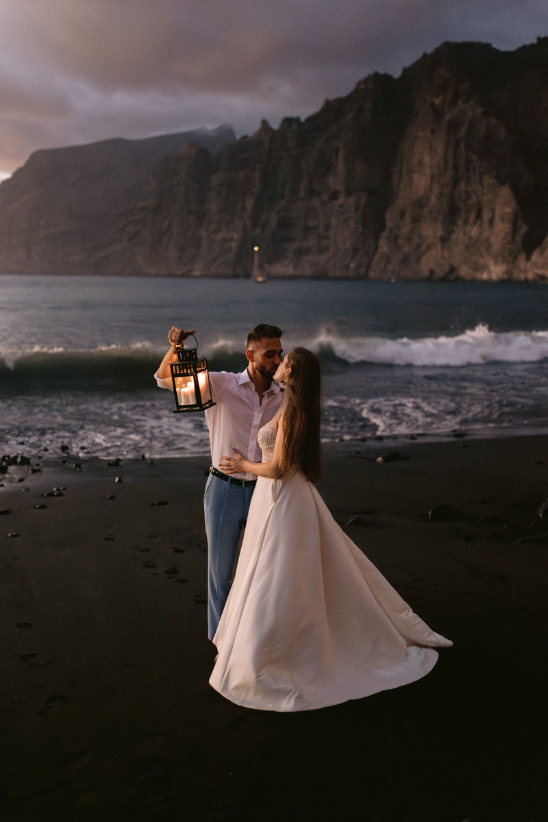 Wedding photoshoot of a couple kissing at dusk in front of Los Gigantes in Tenerife on the beach, holding a lantern